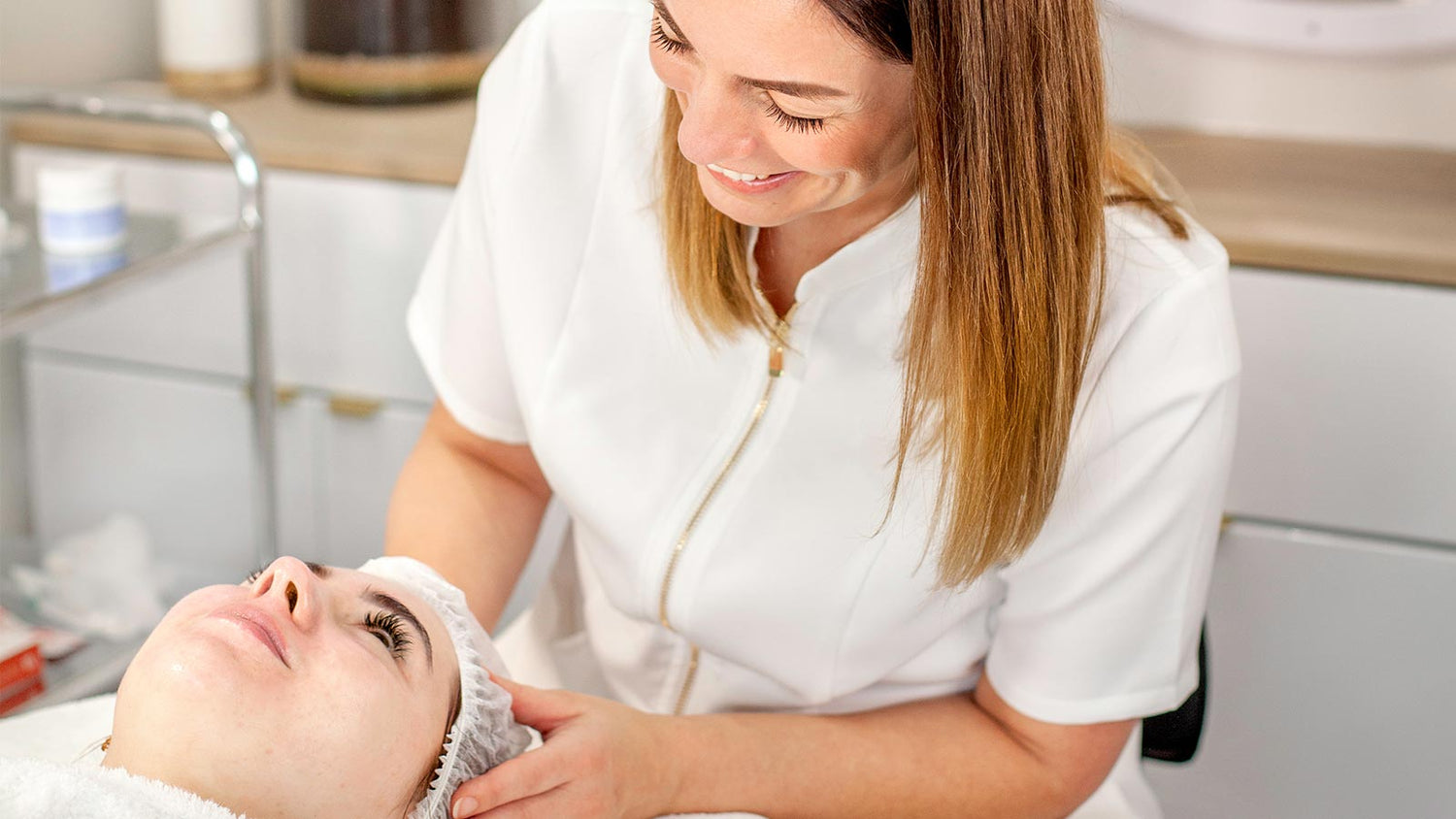 Leah, a skincare specialist, smiling while giving a facial treatment to a client lying on a treatment bed.