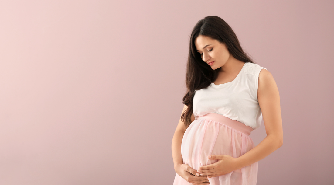 A pregnant woman wearing a white top and pink skirt gently holding her bump while looking down with a calm expression.