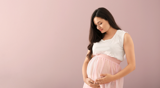 A pregnant woman wearing a white top and pink skirt gently holding her bump while looking down with a calm expression.