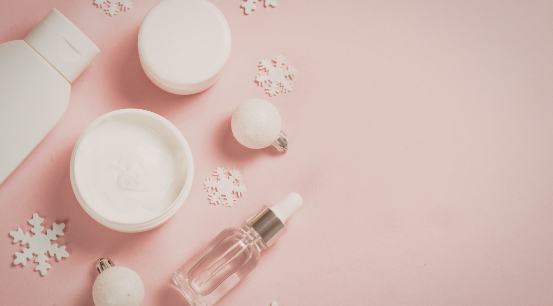 Winter-themed flat lay of skincare products including moisturiser, serum and lotion on a pink background with snowflake decorations.